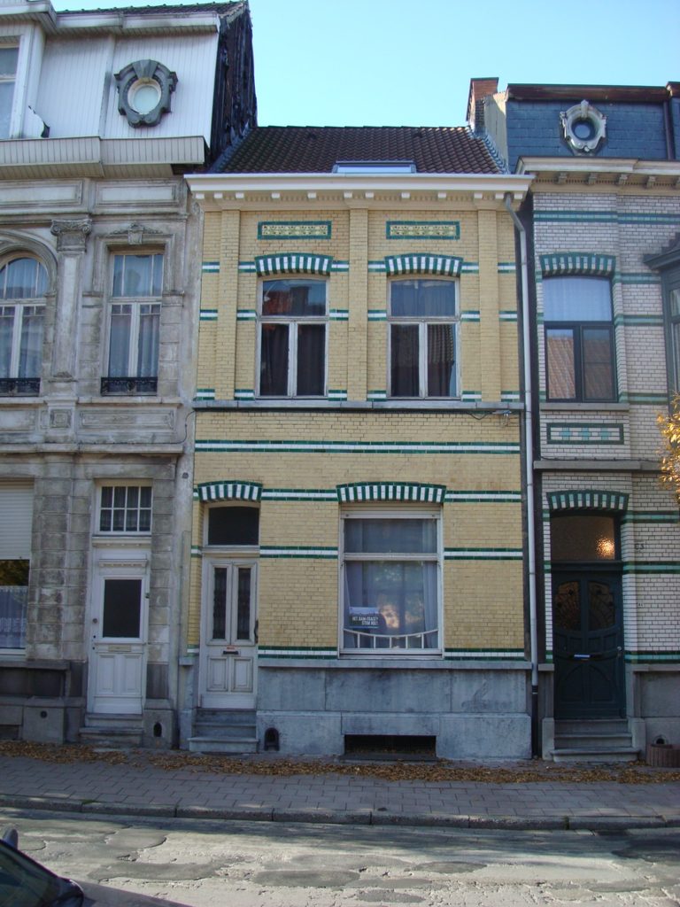 “Front view of a yellow-brick Art Nouveau house between two neighbours: a plain rendered grey facade on the left, whose boundary strip runs with it, and a glazed cream-and-green brick facade on the right, the older of the two neighbours, whose strip was originally the one built and finished first.”