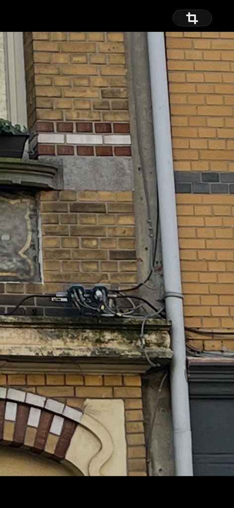 “Close-up of the seam between two facades, showing a tangle of electrical cables and a junction box mounted on the cornice edge, next to a weathered ornamental panel and a red-and-white brick band.”