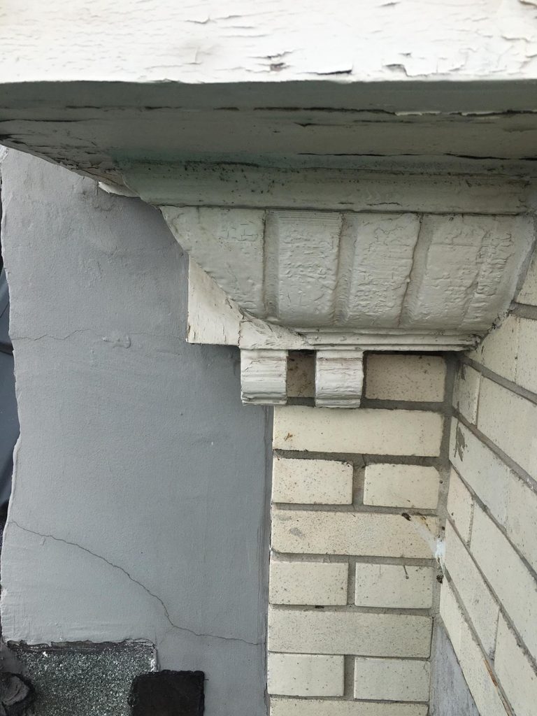 “Close-up of a peeling, painted cornice with decorative brackets: the grey rendered strip belonging to the yellow-brick facade, projecting outward and overhanging the neighbouring cream glazed-brick facade below it.”