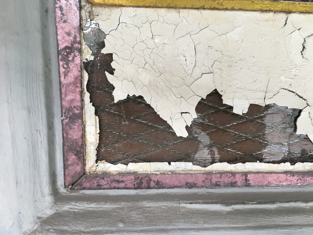 Close-up of a damaged glass panel corner, showing the layered construction beneath cracked paint: diamond-pattern reinforced glass, a bed of putty, and a frame edge finished in pink with a metallic sheen.