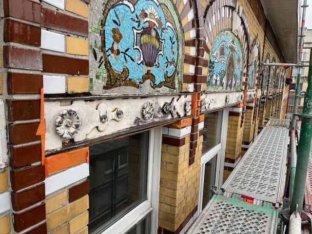 Close-up of a mosaic frieze above the windows during restoration, showing the lost Venetian green glas teserae and the exposed, weathered ornamental cornice with scaffold boards visible in the foreground.