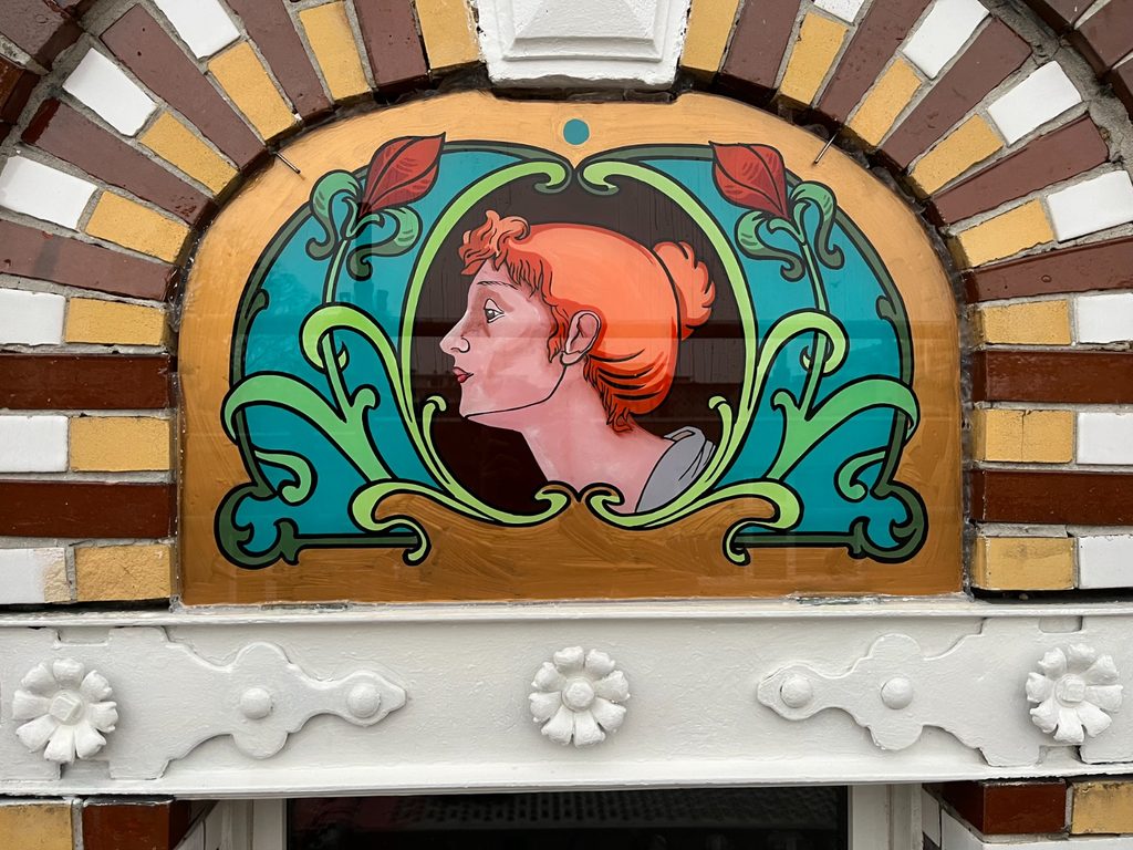 Finished hand-painted glass panel with an arched top, showing a woman's profile with red hair against a teal background, framed by green scrollwork and white flowers with red leaves, set into a brick archway.