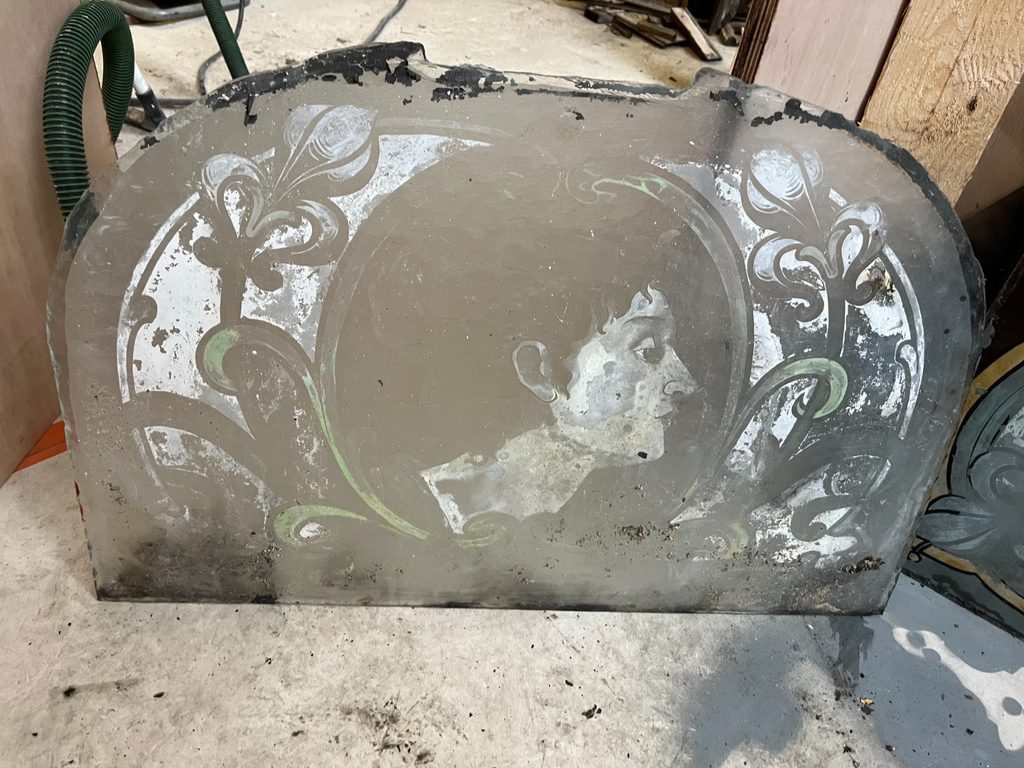 Weathered glass panel with an arched top, showing a woman's face in profile faintly visible at the centre, surrounded by white and partially green Art Nouveau floral scrollwork.
