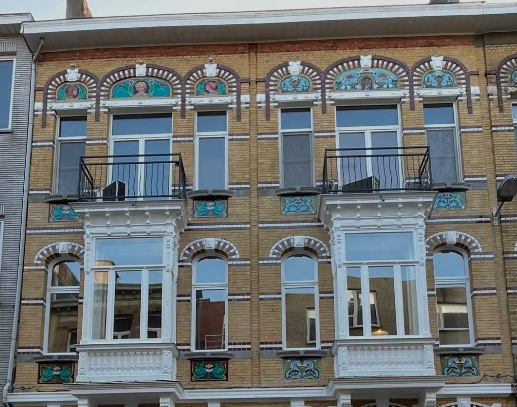 Art Nouveau townhouse facade with bay-window balconies, the window lintels decorated with mosaic panels in blue, green, and terracotta, which arent restored alternating with polychrome brick arches on the right side , and on the left you see the new painted glass plates.