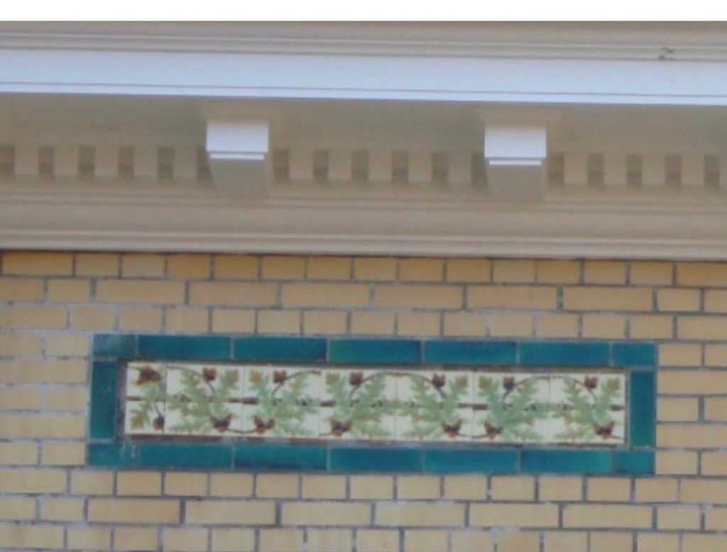 Yellow brick facade detail showing a green-bordered ceramic tile band with an oak leaf motif.