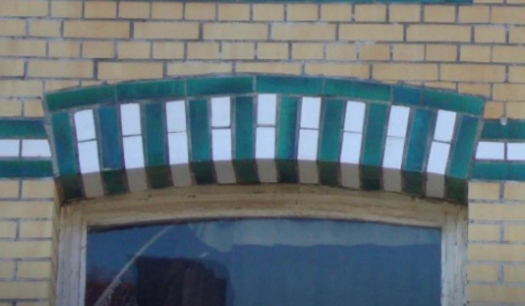 Arched window opening framed by alternating green and white glazed brick voussoirs, set into a yellow brick facade.