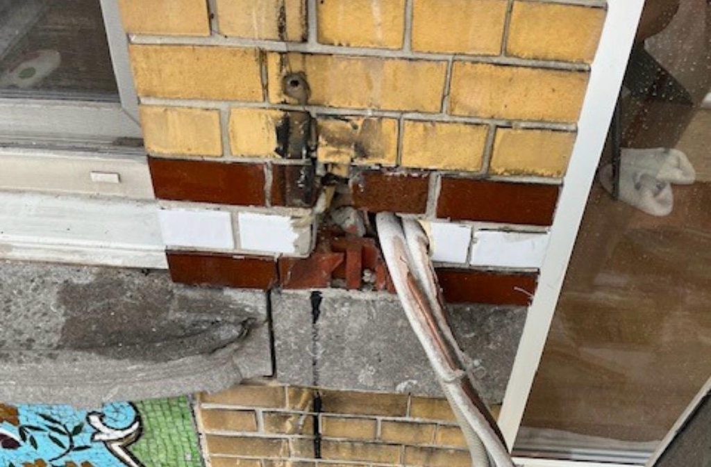 Close-up of a repair in progress on a glazed brick facade, showing a white conduit pipe running down between rows of brown, white, and yellow glazed brick, with a fragment of turquoise mosaic visible at the edge.