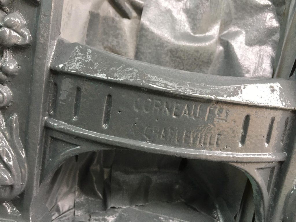 Close-up of the curved cross bar of a cast-iron boot scraper, freshly painted in glossy grey. The raised lettering "CORNEAU Frs CHARLEVILLE" is clearly legible, with narrow vertical slots on either side.