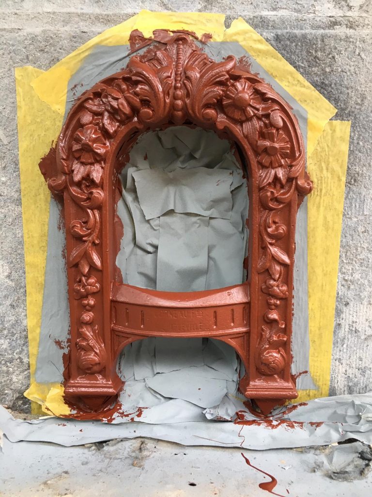 Cast-iron boot scraper in an arched frame, set in a stone wall and painted red-brown, with masking tape and paper protecting the surrounding stone. The arch is covered in relief of acanthus leaves, scrolls and daisy-like flowers, with a beaded ridge at the top and the name on the curved scraper bar.