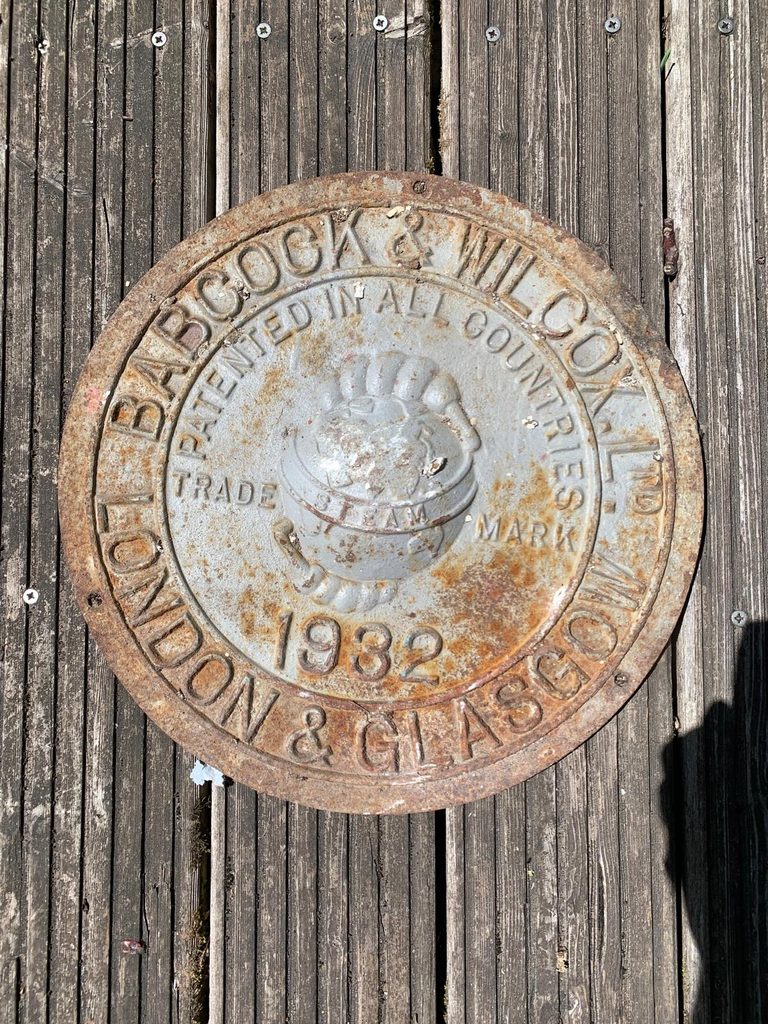 Round cast-iron plate with raised lettering reading "Babcock & Wilcox Ltd, London & Glasgow, 1932", with a steam mark emblem in the centre and the words "Patented in all countries". Rusty, with flaking grey paint, lying on weathered wooden decking.